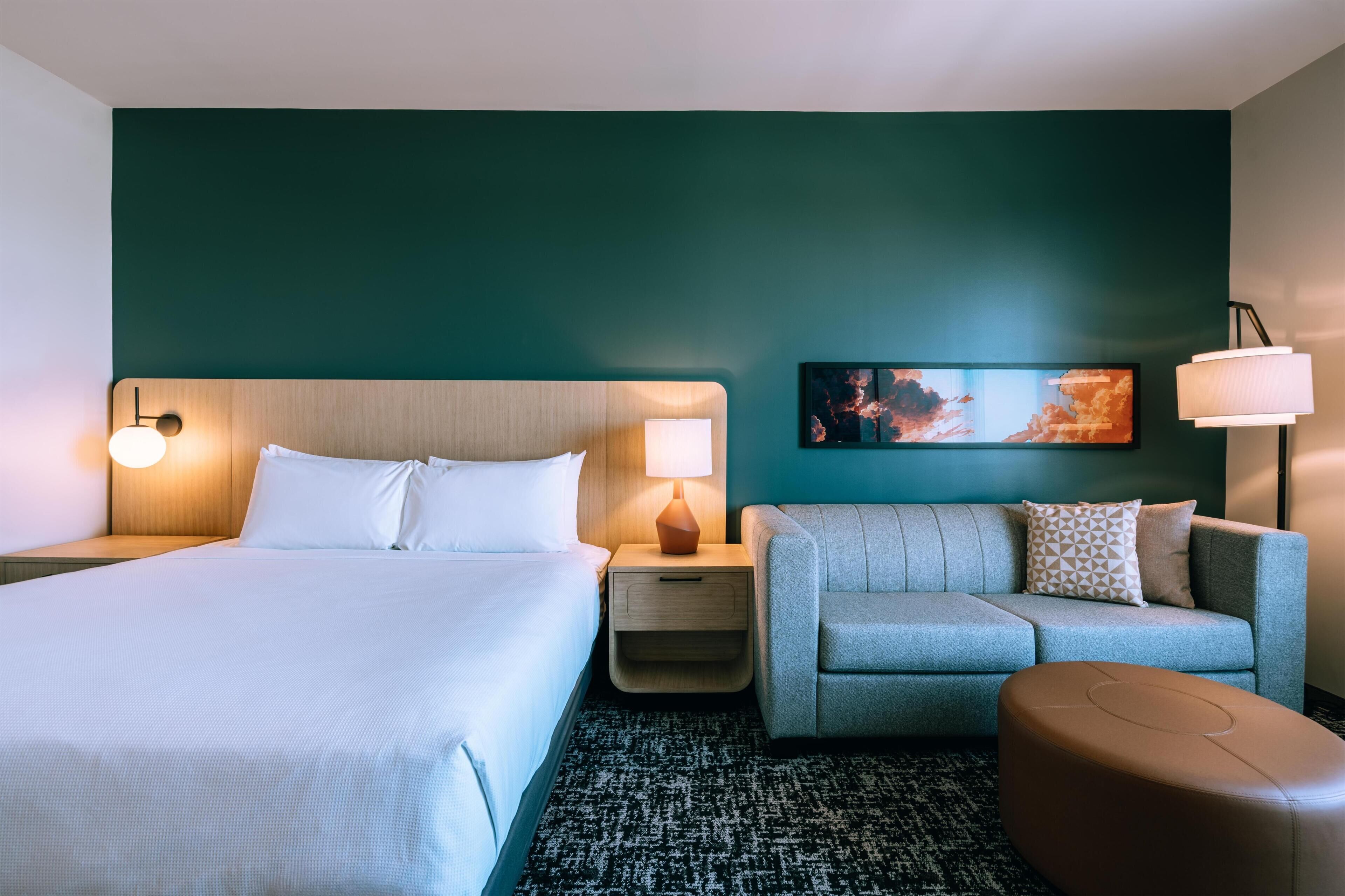 Habitación de hotel con cama, sofá, mesita de noche, lámpara y un cuadro en la pared.