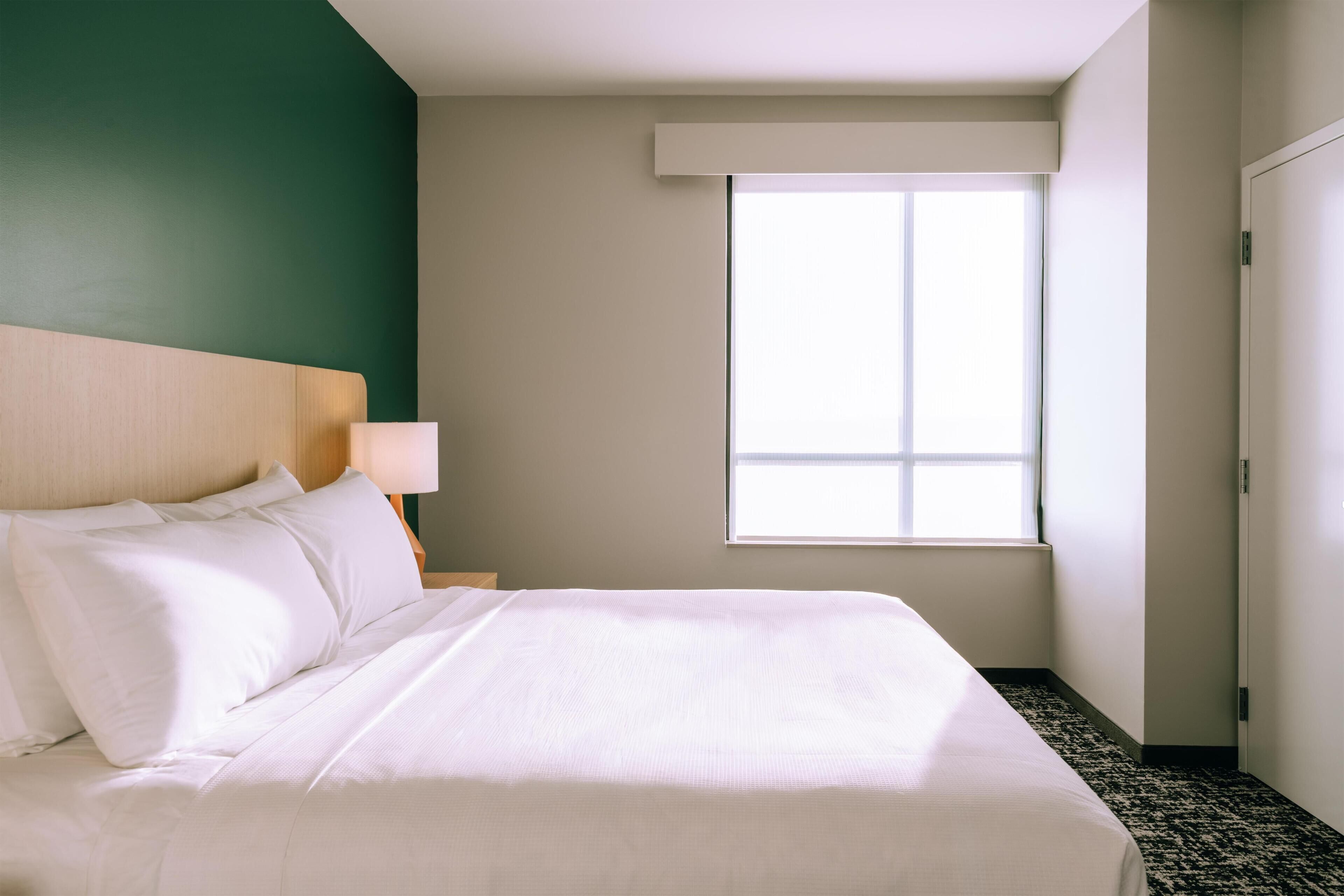 Quarto de hotel com cama, mesa de cabeceira, abajur, janela e porta.
