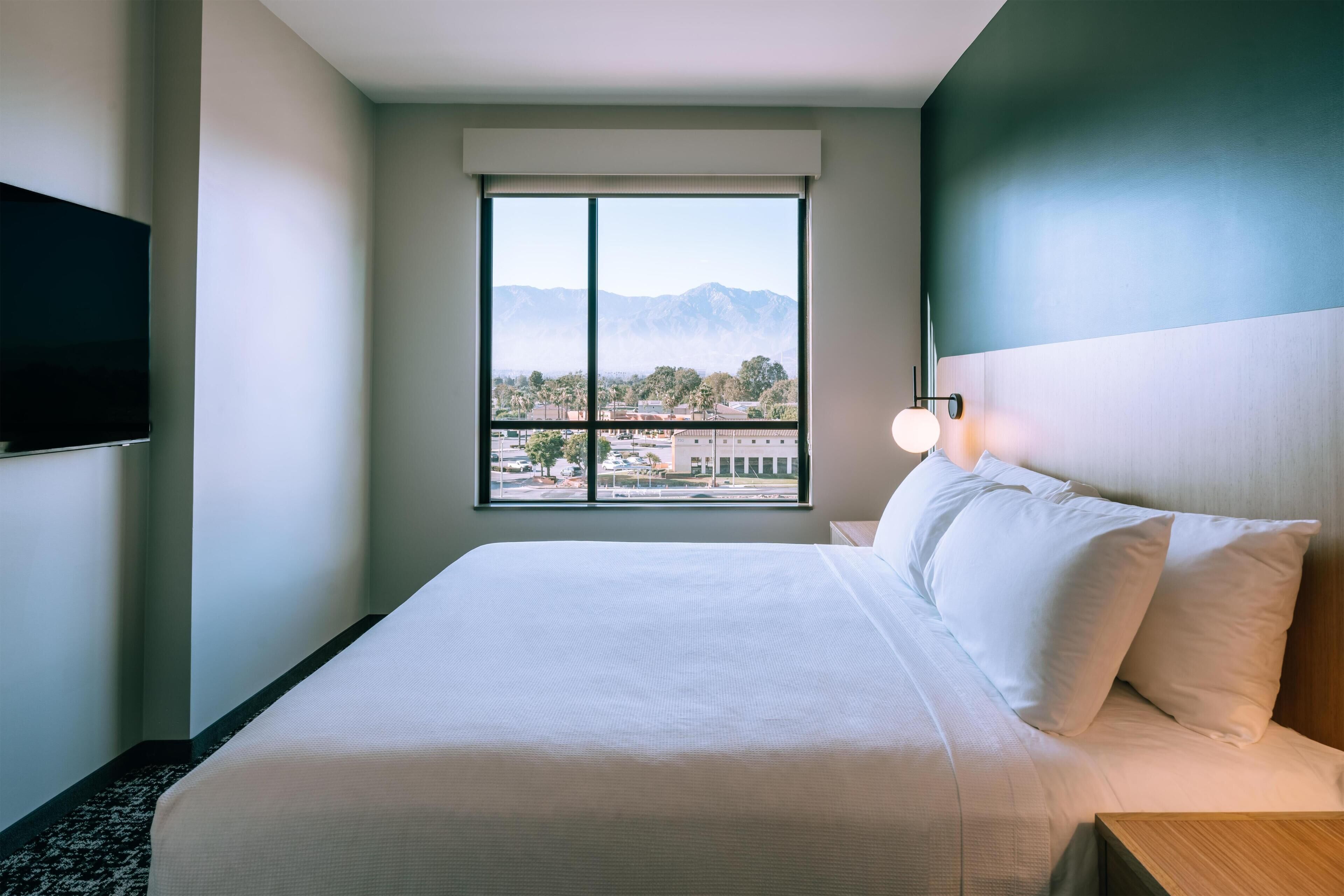 A hotel room with a large bed, a flat-screen TV mounted on the wall, and a window with a mountain view.