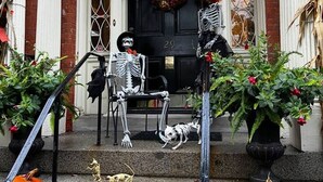 A building entrance with a black door, a skeleton figure sitting on the steps, and a skeleton figure standing on the porch.