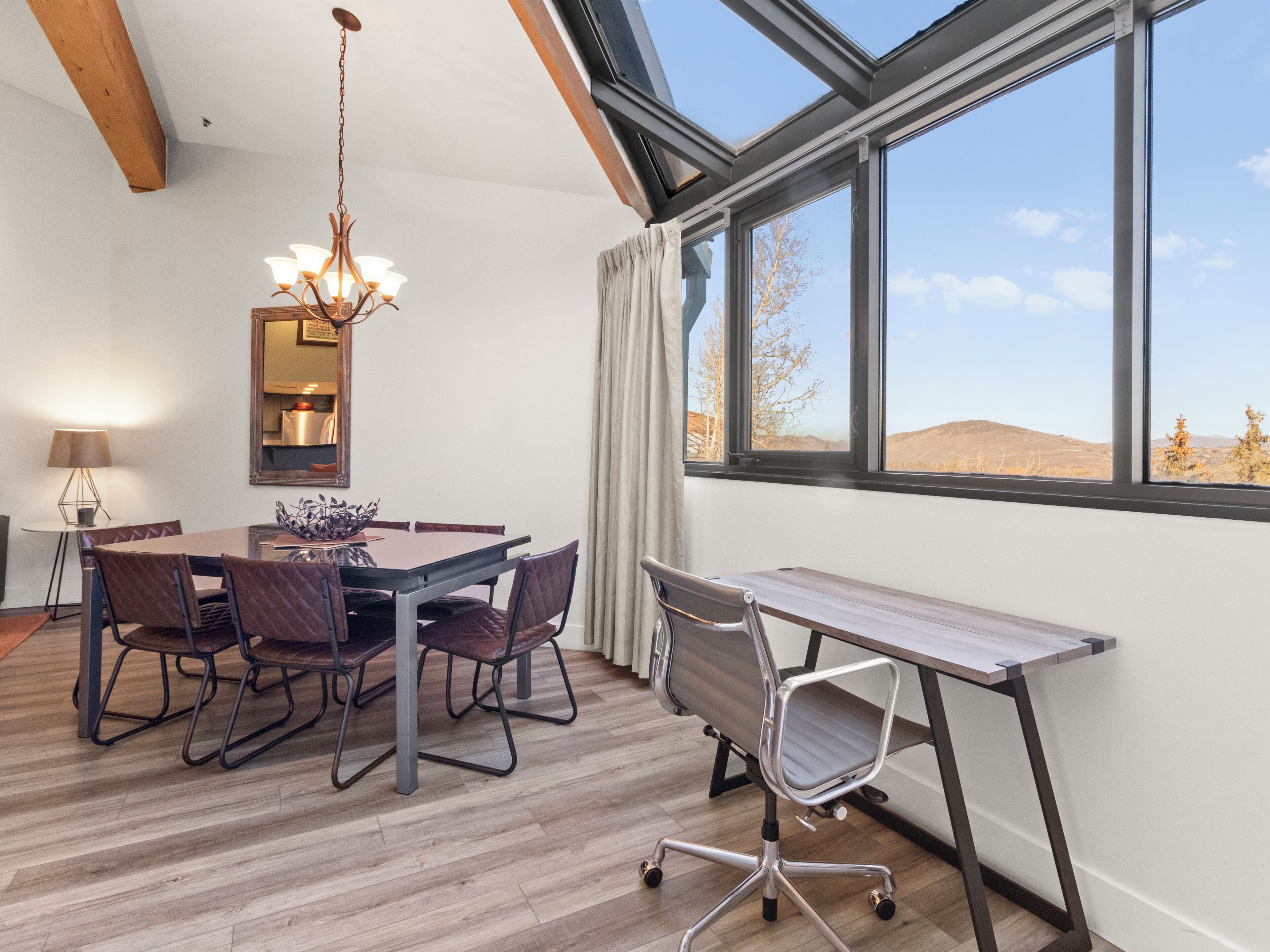 A modern dining area with a wooden table, cushioned chairs, a chandelier, and a large window offering a view of the landscape.