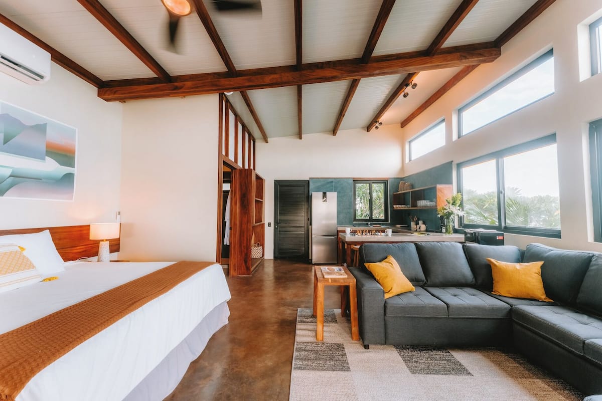 A modern hotel room with a bed, a sofa, a dining area, and a kitchen.