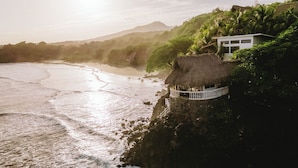 A coastal scene with a thatched-roof structure on a cliff, overlooking a beach and a mountain in the background. - Punta Mango Villas (Jucuaran)