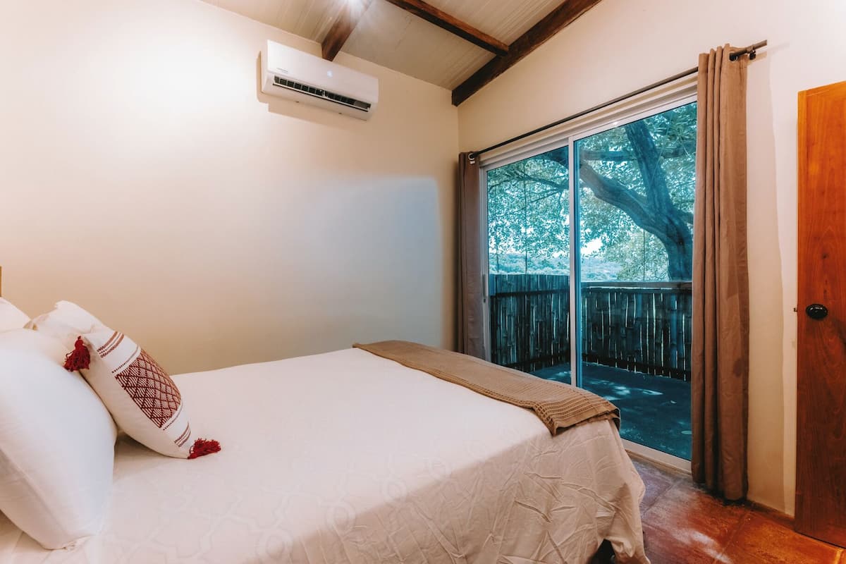 A bedroom with a bed, an air conditioner, a window with curtains, and a view of trees.