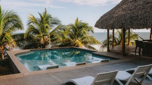 A swimming pool with a view of the ocean, surrounded by palm trees and a thatched roof structure. - Punta Mango Villas (Jucuaran)