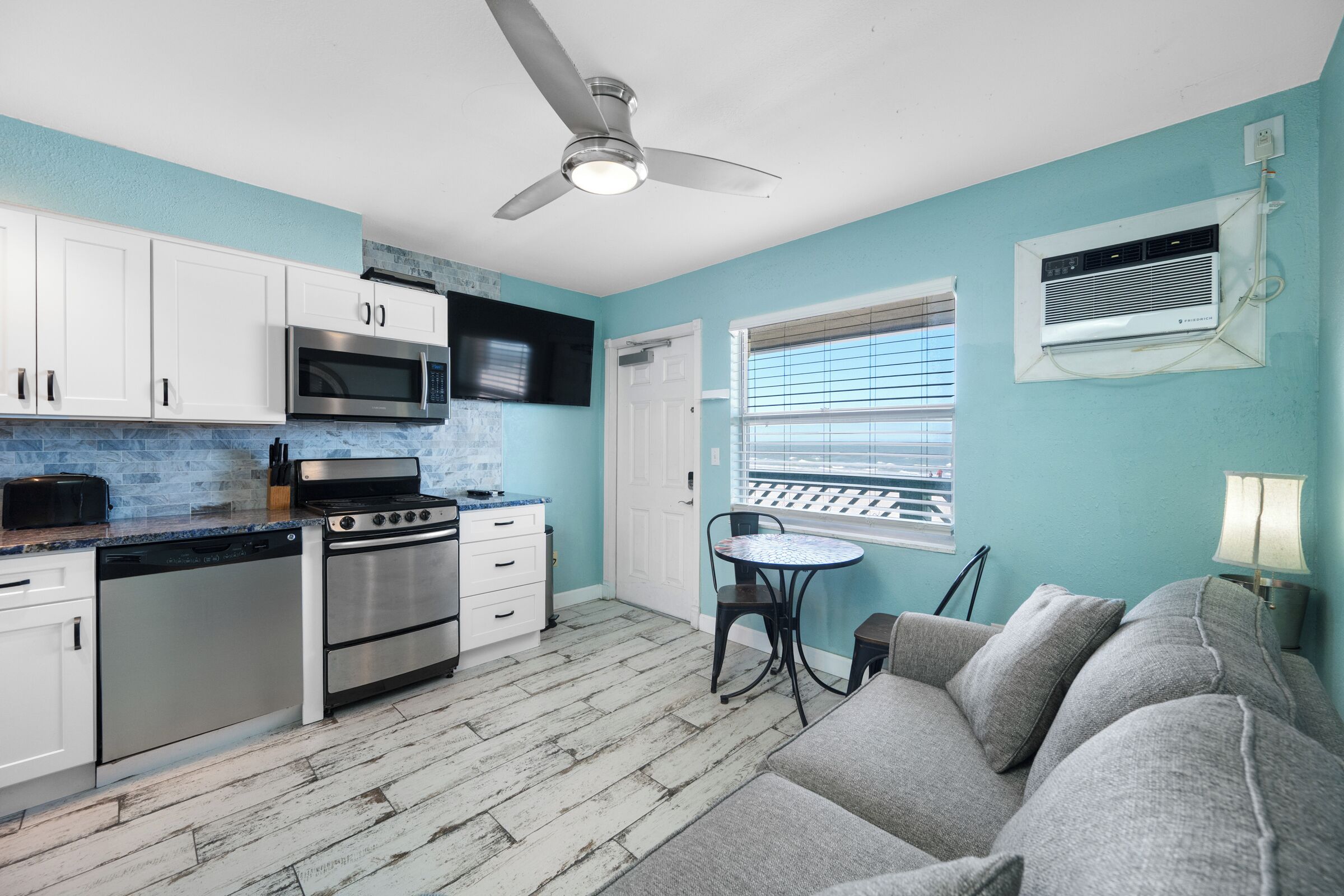 A compact kitchen with a microwave, stove, and dishwasher, a ceiling fan, a window with blinds, and a gray couch.