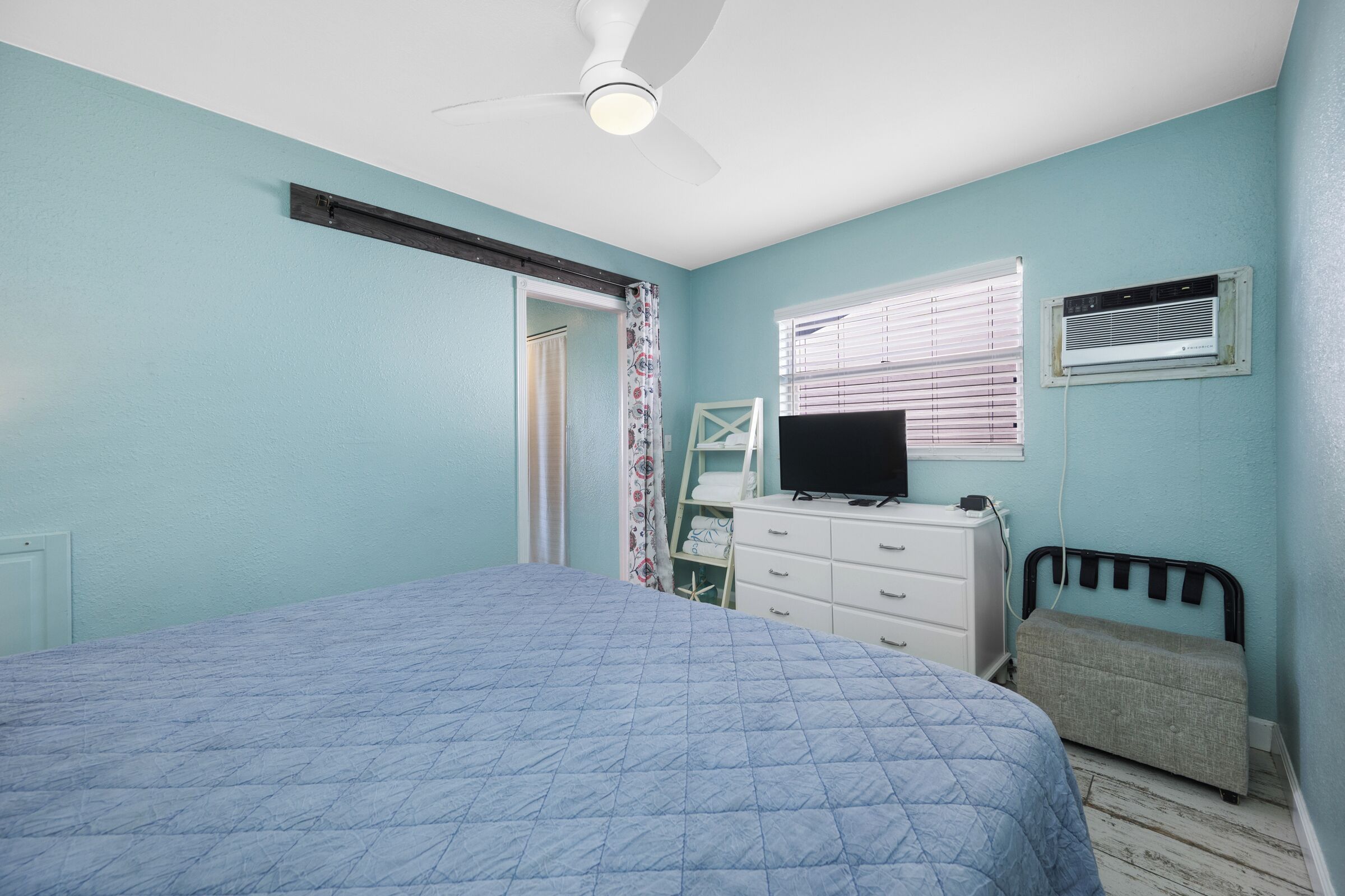 Une chambre à coucher comprenant un lit, un ventilateur de plafond, une télévision, une commode et une fenêtre avec des stores.