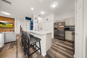 A modern kitchen with a central island, bar stools, a refrigerator, and a dining area with a sofa and a TV. - 4720 HH - Fiesta Key Grandeur (Kissimmee)