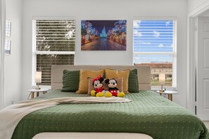 A bedroom with a green bedspread, two Mickey Mouse pillows, a framed picture of a nighttime cityscape, and a window with blinds. - 4720 HH - Fiesta Key Grandeur (Kissimmee)