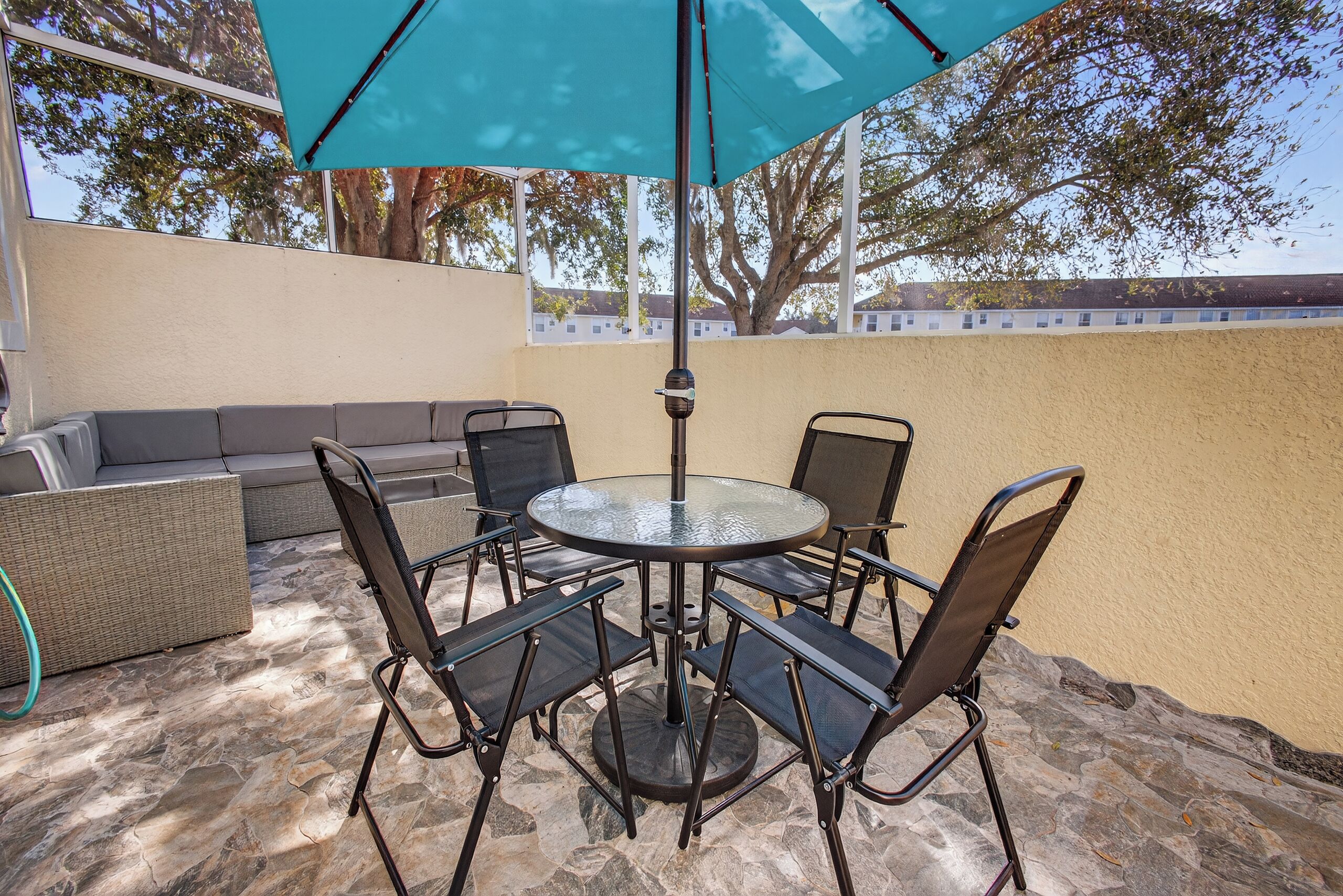 Une terrasse extérieure avec une table ronde en verre et quatre chaises, un canapé en osier et un grand parasol bleu.