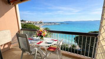 A balcony with a table set for two, overlooking a coastal view with a marina and buildings in the distance.