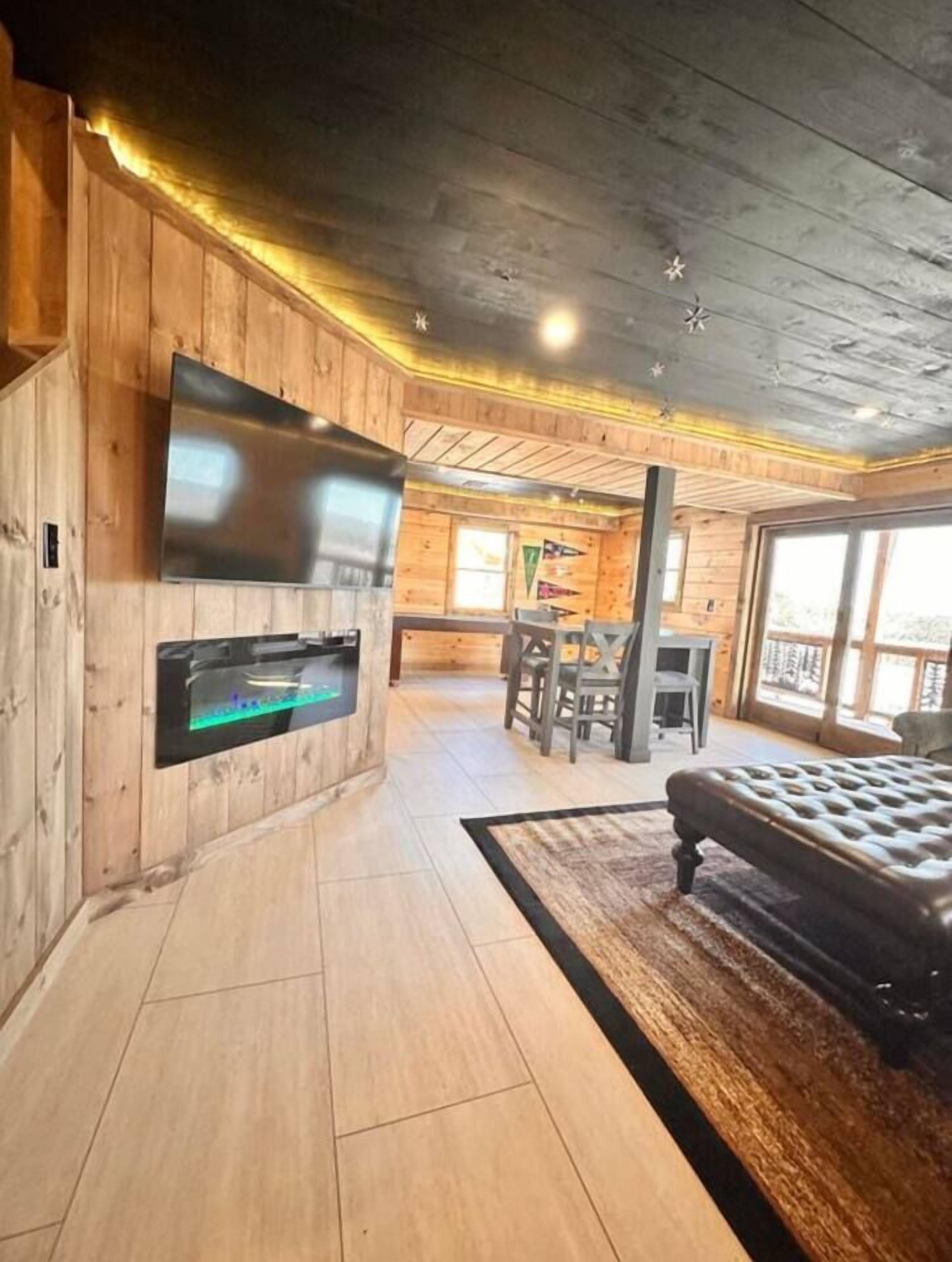 A wooden interior with a flat-screen TV mounted on the wall, a seating area with a table and chairs, and a large window providing natural light.