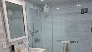 Un baño moderno con ducha envuelta en vidrio, paredes con azulejos blancos, un lavamanos blanco y un espejo encima, y un inodoro blanco.