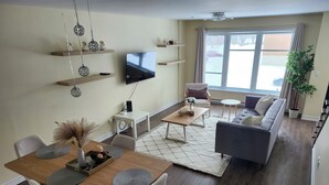 A modern living room with a dining area, a flat-screen TV mounted on the wall, and a large window providing a view of the outside. - Modern 2BR Furnished Home near DIX30 & Downtown Montreal— Monthly Stays Welcome (Longueuil)