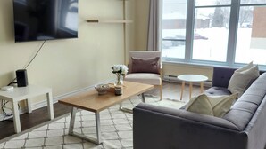 A modern living room with a grey sofa, a coffee table, a TV mounted on the wall, and a large window with a view of the outside. - Modern 2BR Furnished Home near DIX30 & Downtown Montreal— Monthly Stays Welcome (Longueuil)