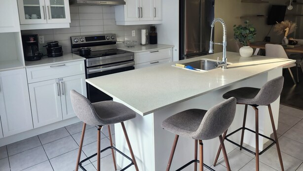 A modern kitchen with white cabinetry, stainless steel appliances, and a central island with bar stools. - Modern 2BR Furnished Home near DIX30 & Downtown Montreal— Monthly Stays Welcome (Longueuil)