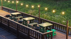 A deck with picnic tables and a grill, surrounded by a wooden railing and string lights. - Gangneung Trip Spa Pension (Gangneung)