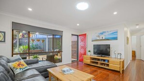 A modern living room with a sofa, coffee table, TV, and a view of the outdoors through large windows. - Tranquility At Lighthouse Beach (Port Macquarie)
