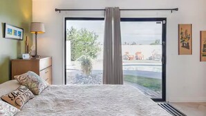 A bedroom with a bed, a nightstand, a lamp, a sliding glass door leading to a pool area. - Hip 3-bedroom House with AC/Pool/Hot Tub in Vibrant Palm Springs (Palm Springs)