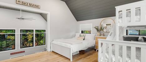 A bedroom with a bed, a crib, a ceiling fan, a window with a view of trees, and a sign on the wall.