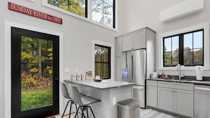 Une cuisine moderne dotée d’un grand îlot central, de tabourets de bar, d’une poubelle en acier inoxydable et offrant une vue sur une forêt grâce à une porte vitrée.