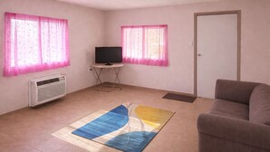 Ein Wohnzimmer mit einem Flachbildfernseher auf einem Schrank, einer Couch, einem Teppich und zwei Fenstern mit rosa Vorhängen.