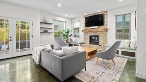 A modern living room with a gray sofa, a coffee table, and a TV mounted on a stone wall. - Charming Coastal Getaway – 5 Min to Boardwalk! (Santa Cruz)