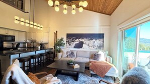 A modern living room with a sofa, coffee table, and chairs, a kitchen with bar stools, and a large painting. - Summit View Collection |Ski-In ·Hot Tub ·Sleeps 14 (Kimberley)