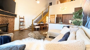 A spacious living area with a fireplace, a staircase leading to an upper level, a kitchen, and a cozy seating area with a sofa and a coffee table. - Summit View Collection |Ski-In ·Hot Tub ·Sleeps 14 (Kimberley)