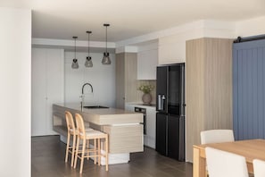 A modern kitchen with a central island, a black refrigerator, and a dining area with wooden chairs. - Luxury Beachfront Burleigh Heads (Burleigh Heads)