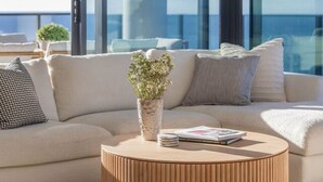 A modern living room with a round wooden coffee table, a beige sofa with patterned cushions, and a vase with flowers. Large windows provide a view of the sky and a distant building. - Luxury Beachfront Burleigh Heads (Burleigh Heads)