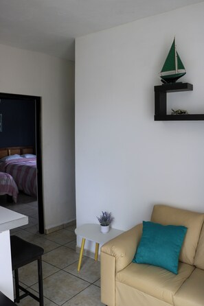A room with a beige sofa, a small white table with a plant, a bed with pink bedding, and a green sailboat model on a black shelf.