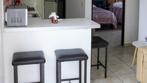 A compact kitchen with a microwave, stove, and sink. There are two black stools and a small table. A bed with pink bedding is visible through an open door. - Apartment No. 2, perfectly located with swimming pool (pets welcome) (Ciudad Madero)
