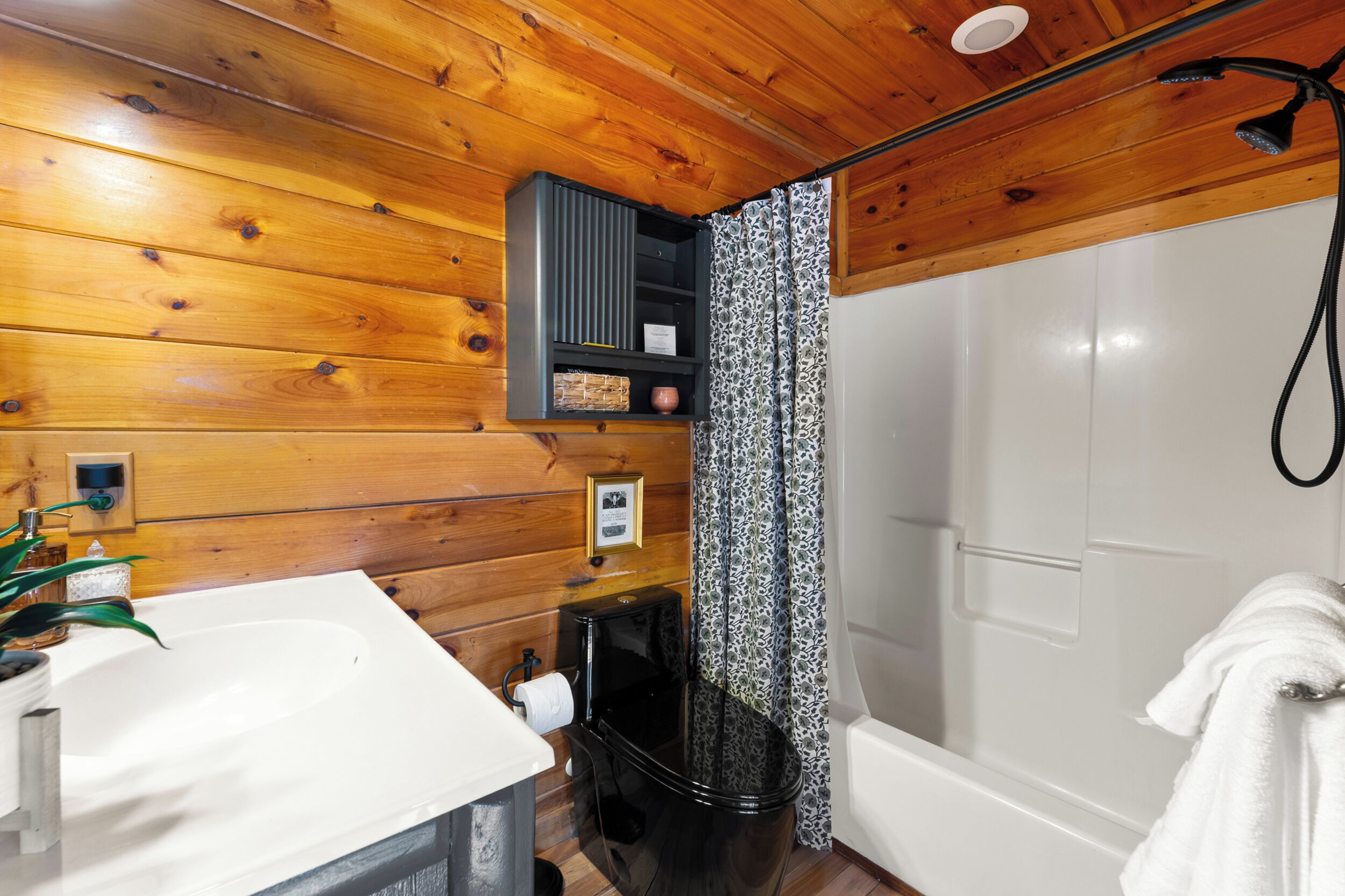 Ein Badezimmer einer Holzhütte mit Waschbecken in Weiß, Toilette in Schwarz und einer Duschkabine.