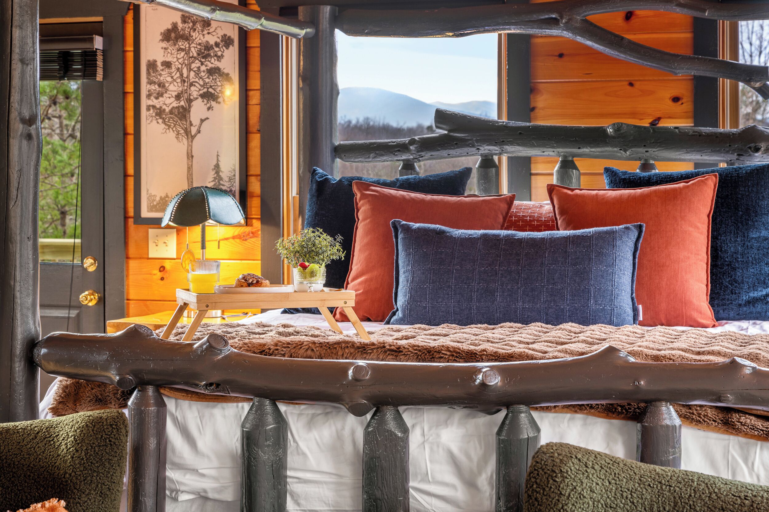 Ein rustikales Schlafzimmer mit einem Holzbett, Blick ins Freie durch ein Fenster und einem kleinen Tisch mit Lampe und Blumen.