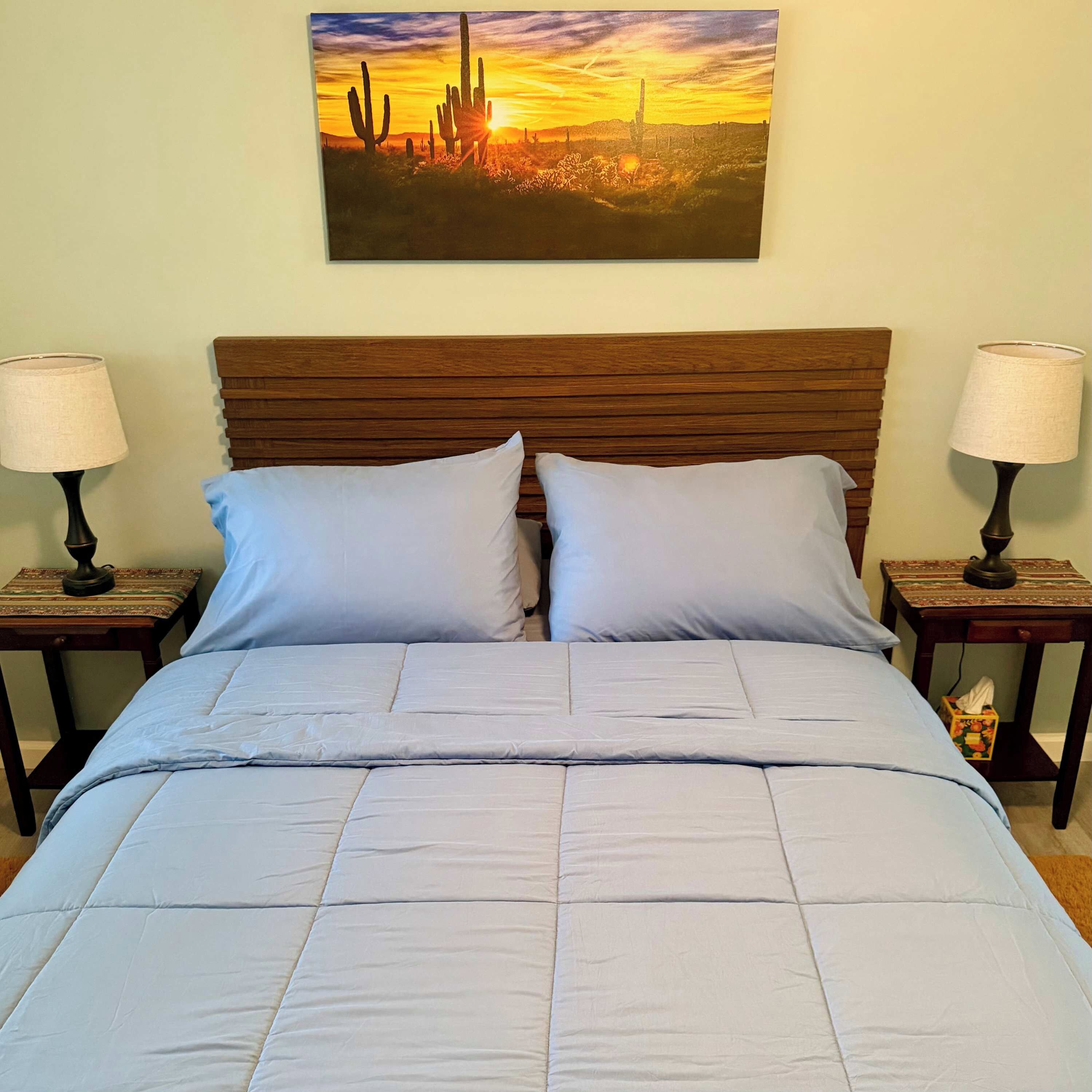 A bed with a wooden headboard, two nightstands with lamps, and a painting of a desert landscape on the wall.