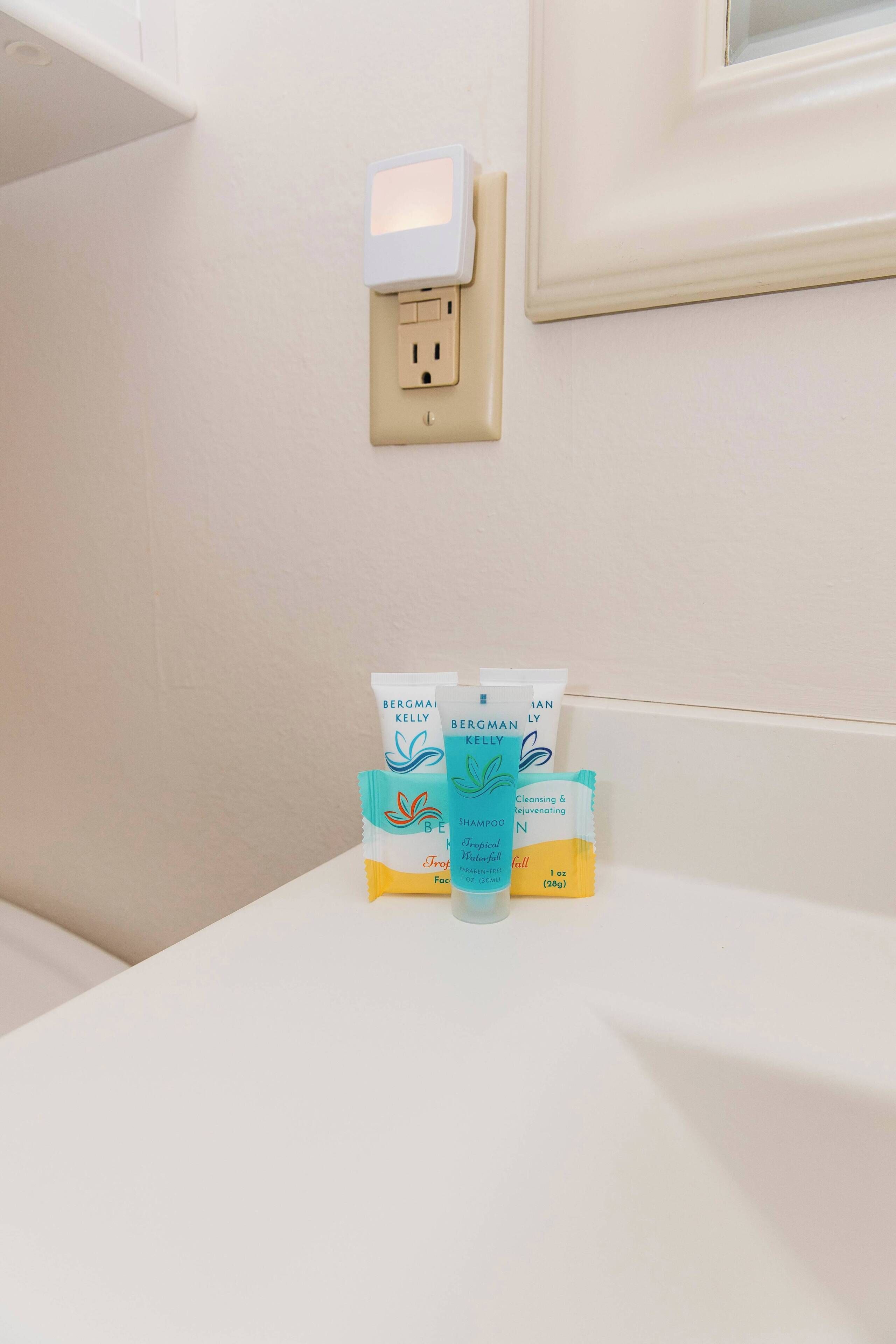 A bathroom with a white sink, a white electrical outlet, and two Bergman Kelly shampoo and conditioner tubes on the counter.