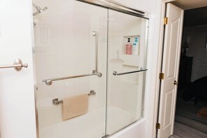 Une douche fermée par des parois en verre, avec un porte-serviettes, une porte dotée d’une poignée en laiton et une petite étagère contenant des articles de toilette.