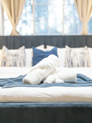 A neatly made bed with towels and a blue blanket, a dark headboard, and a window with curtains. - Ballantyne, Private Yard, Work Desk & 1 Gig WiFi (Charlotte)