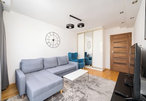 A modern living room with a grey sectional sofa, a white coffee table, and a TV on a black stand. There is a large mirror on the wall, a wooden door, and a blue chair. - Warszawa Wola Center 3-Bedroom Apartment (Warszawa)