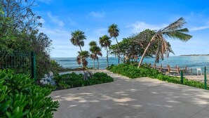 Ein gepflasterter Weg führt zu einem Strand mit Palmen und einem Zaun.