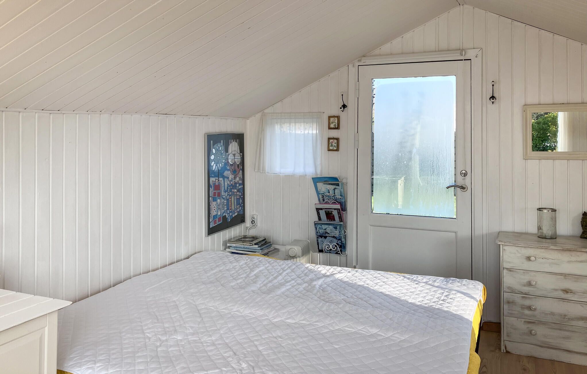 A bedroom with a bed, a dresser, a door with a window, and a wall-mounted picture.