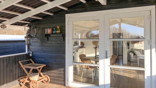 A covered outdoor area with a wooden deck, a wicker cart, and a glass door leading to an interior space with a table and chairs.