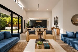 A modern living room with a large window, a dining area, and a kitchen in the background.