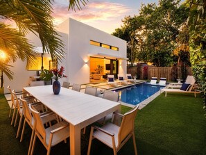 A modern outdoor pool area with a dining table and chairs, surrounded by lush greenery.