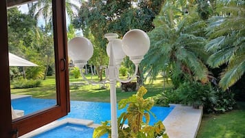 A view from a room with a window overlooking a swimming pool, surrounded by lush greenery and palm trees.