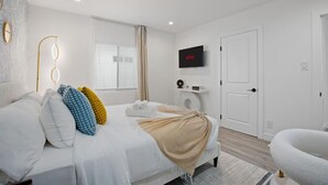 A modern hotel room with a large bed, a flat-screen TV, a bedside table with a lamp, and a view of the street through the window. - Epic Game House w/ Backyard Oasis & Grill in Culver City (Culver City)