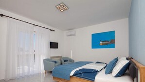 Un dormitorio con una cama de madera, una colcha azul y blanca, una silla azul, una cortina blanca y un cuadro azul en la pared.
