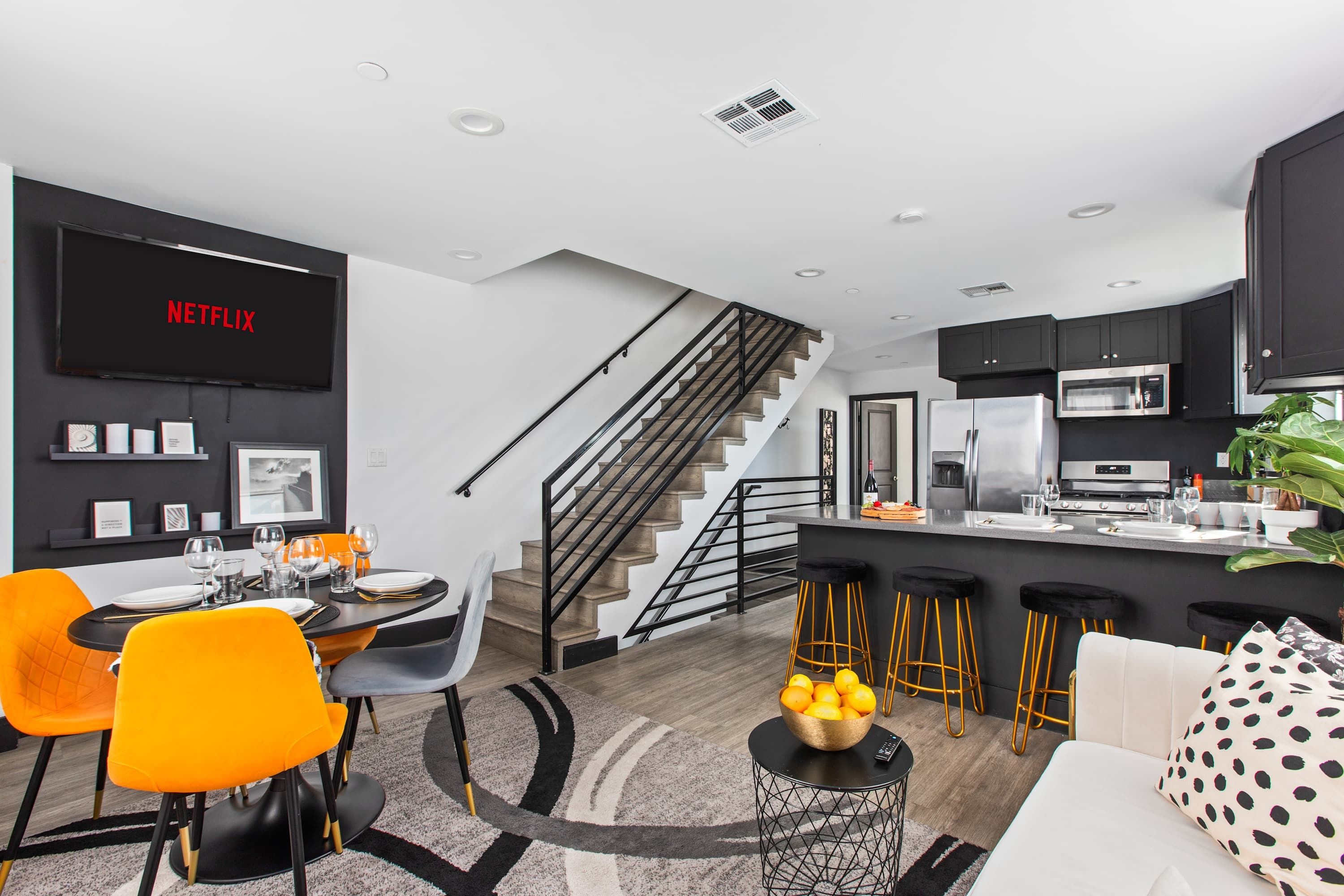 A modern kitchen with a built-in oven, a dining area with orange chairs, and a staircase leading to an upper level.