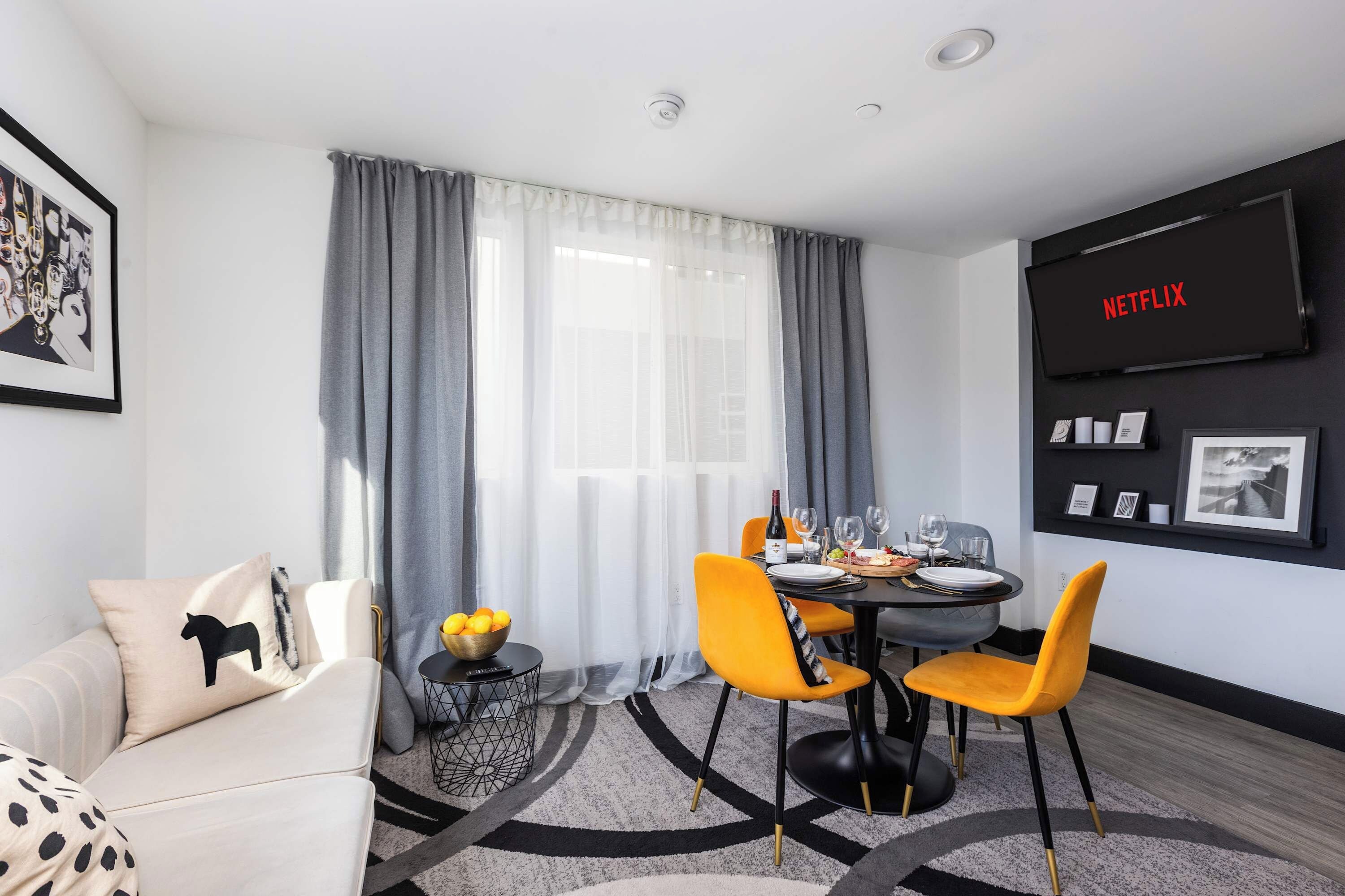 Een moderne woonkamer met een eethoek, een bank, een salontafel met een fruitschaal en een aan de muur bevestigde televisie waarop 'NETFLIX' wordt getoond.